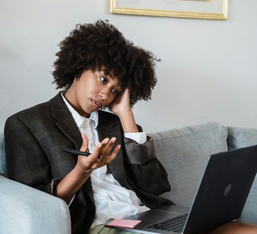 A woman in casual attire sitting on a couch, looking confusedly at her laptop screen.