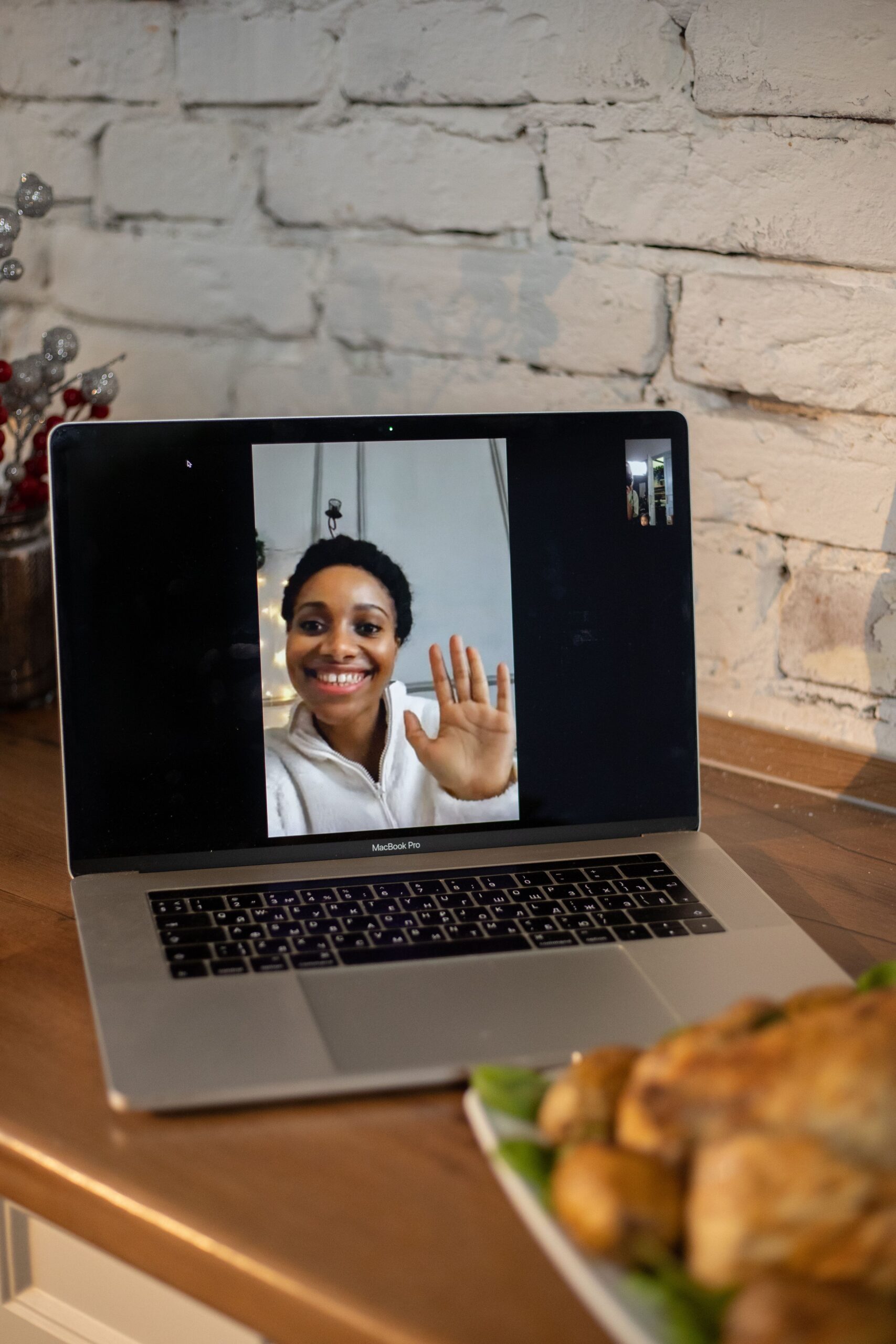 A smiling woman waves during a video call on a laptop, placed on a counter.